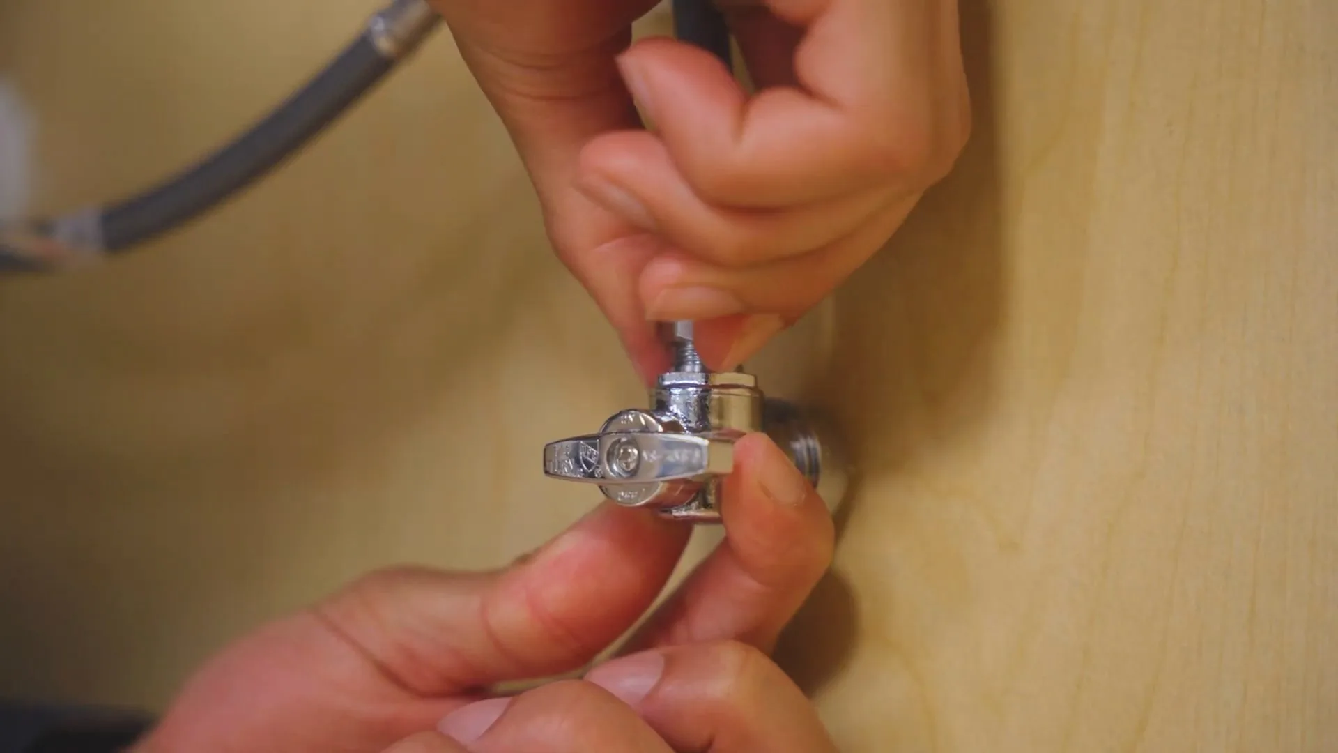 Hands turning a small chrome angle stop valve beneath a cabinet shelf